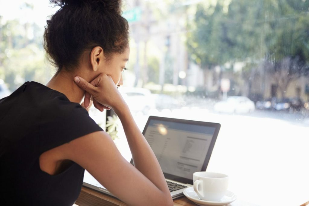 A young BIPOC woman sits at a coffee shop, facing the window, with a cup of coffee and her laptop in front of her, reviewing information on the computer's screen. 