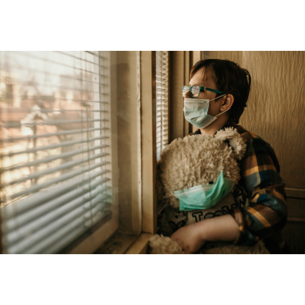 Photo of a woman wearing a face mask looking out the window while holding a teddy bear who is also wearing a face mask.