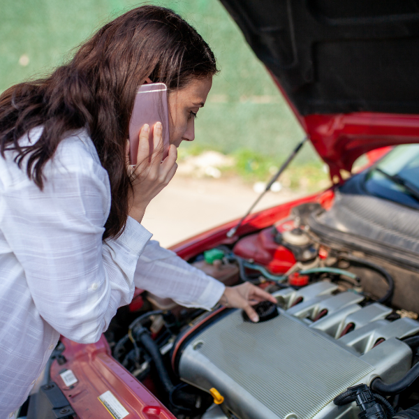 Woman leaned over engine of a car with the hood up talking on the phone with a concerned look on her face.