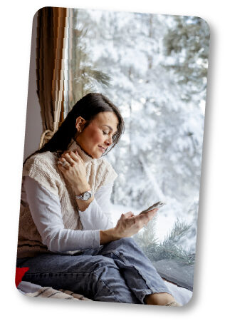 Photo of woman sitting next to a window with a winter landscape while looking at her phone smiling.