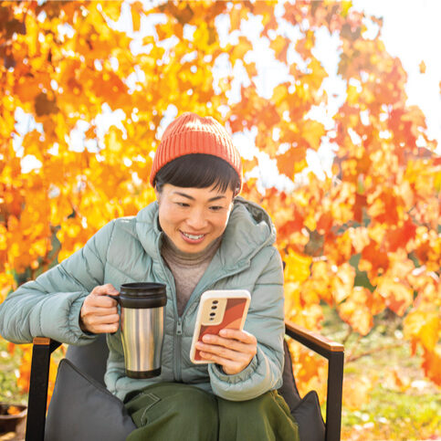 Person sitting outdoors in a chair, holding a travel mug and looking at a smartphone. The person is wearing a light green jacket and an orange knit hat. Bright orange and yellow autumn leaves fill the background in warm sunlight.
