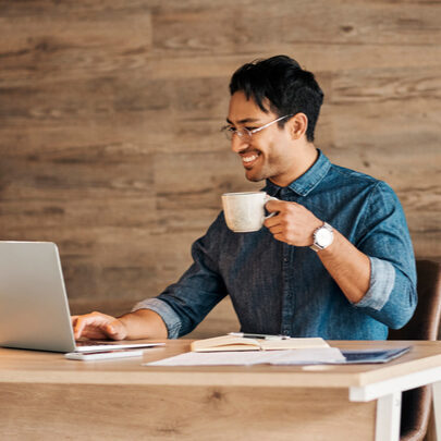 Man working on laptop at desk with a mug in hand while smiling.